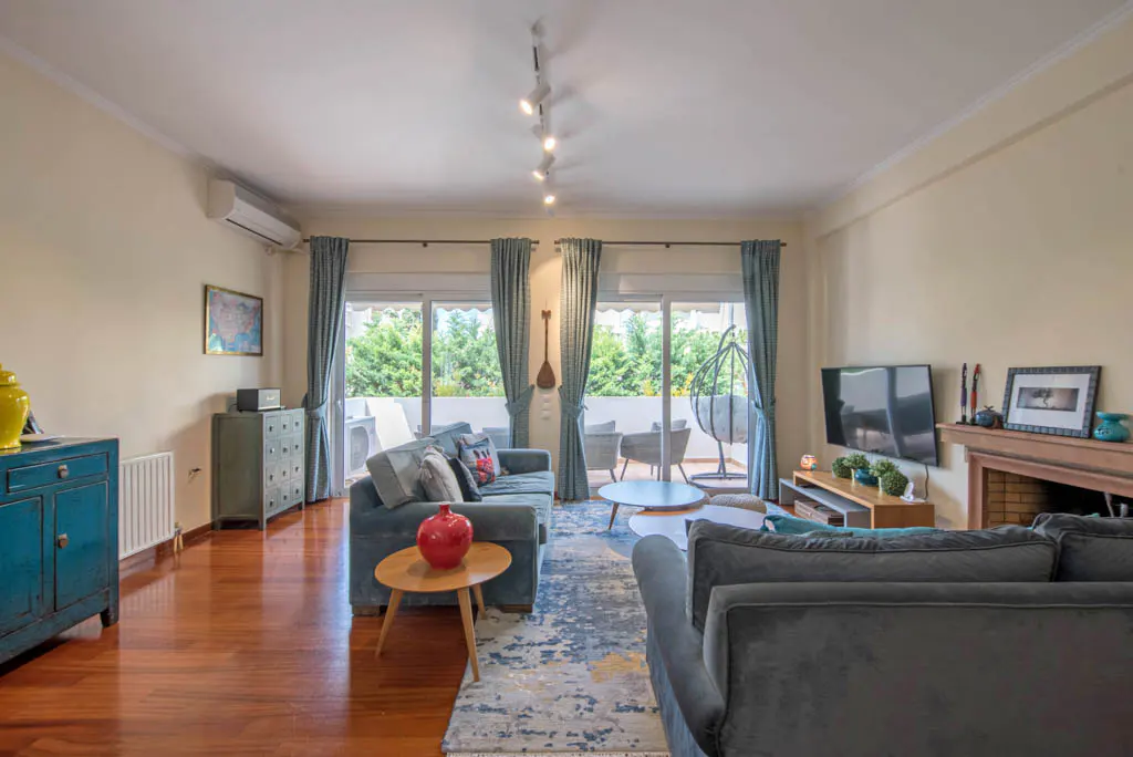 Living room with hardwood floors, blue sofas, and a balcony view. Light walls, blue curtains, and a patterned rug add color.