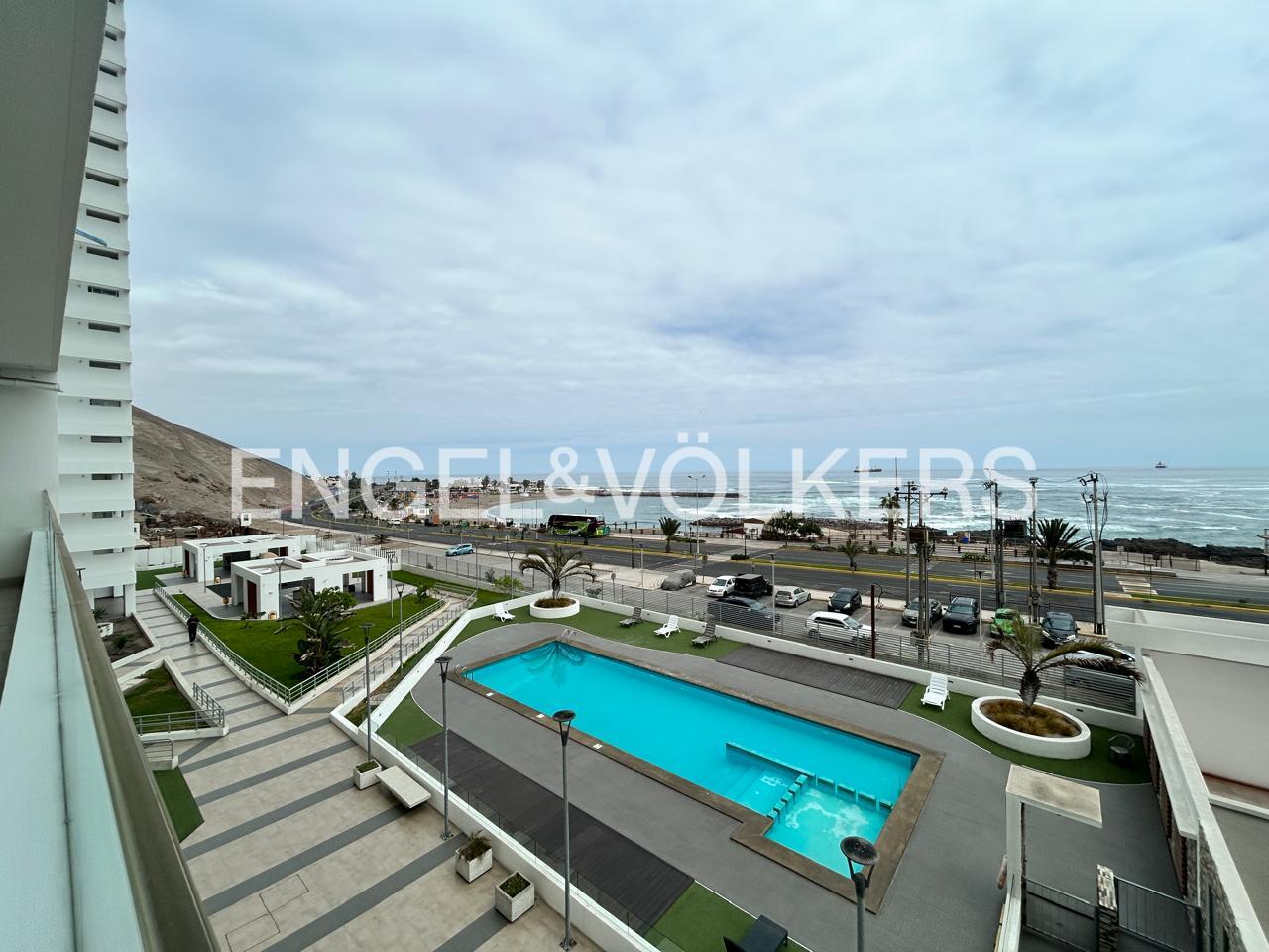 Aerial view of a blue swimming pool, palm trees, and a road with cars, with the ocean in the background on a cloudy day.