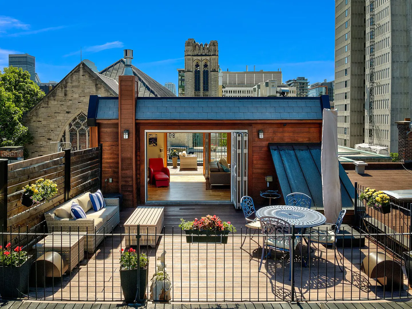Rooftop patio with wood deck, furniture, and open doors to a living room. A church and city buildings are in the background.