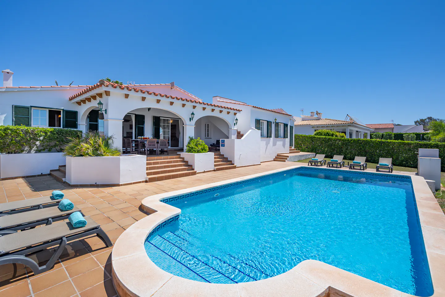 A white villa with a red tile roof overlooks a blue swimming pool and patio with lounge chairs.
