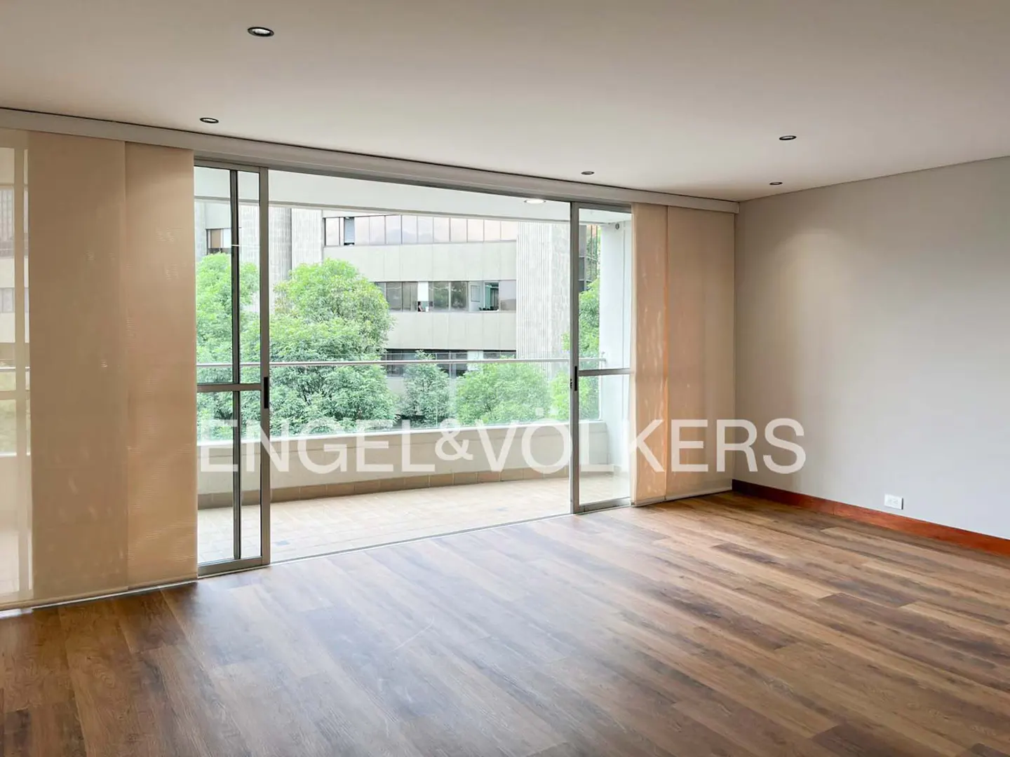 An empty room with hardwood floors and a sliding glass door to a balcony with trees in the background.