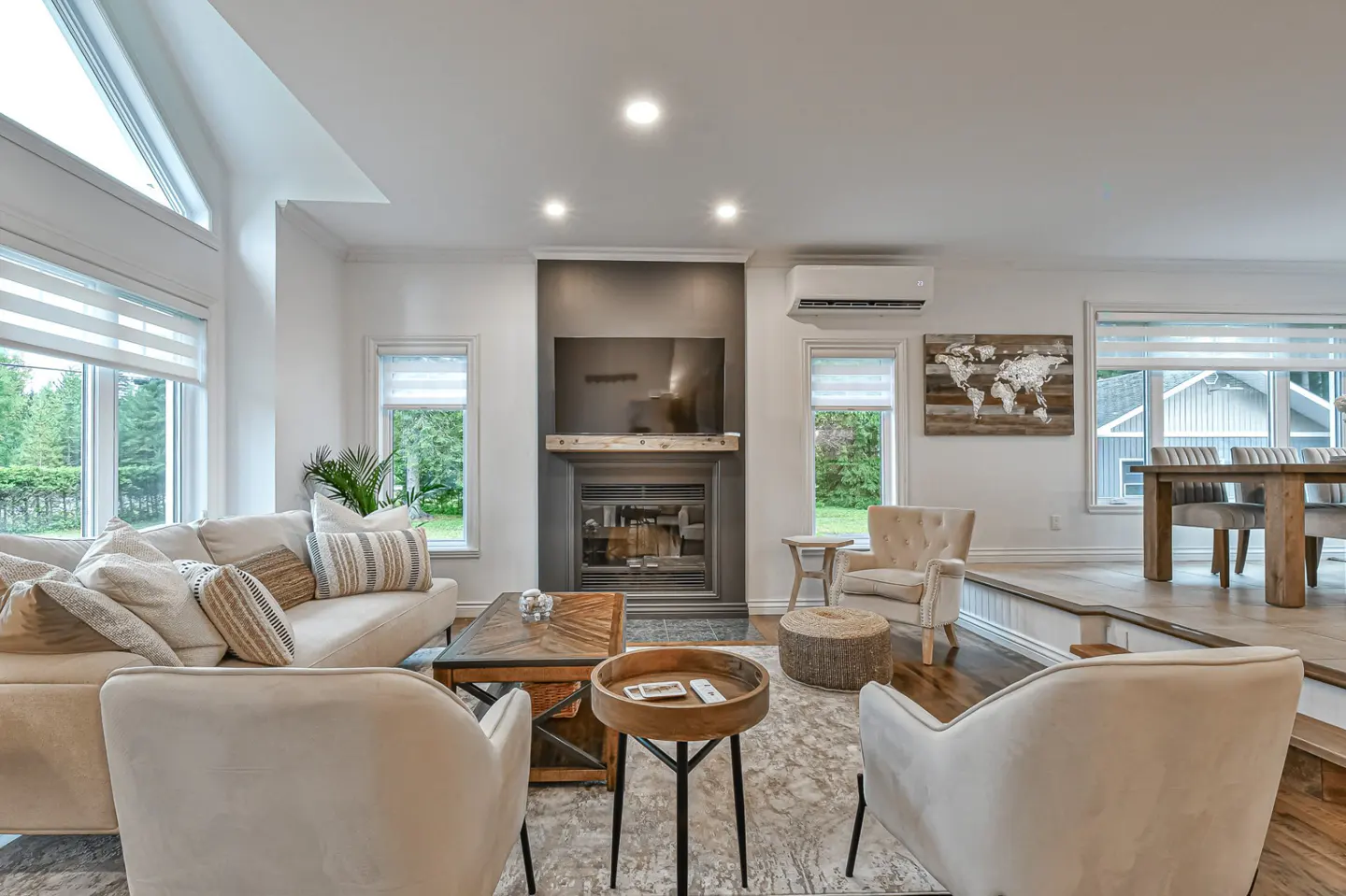 Bright living room with white walls, a fireplace, and large windows. Beige sofa and chairs sit on a gray rug. A wooden dining table is visible in the background.