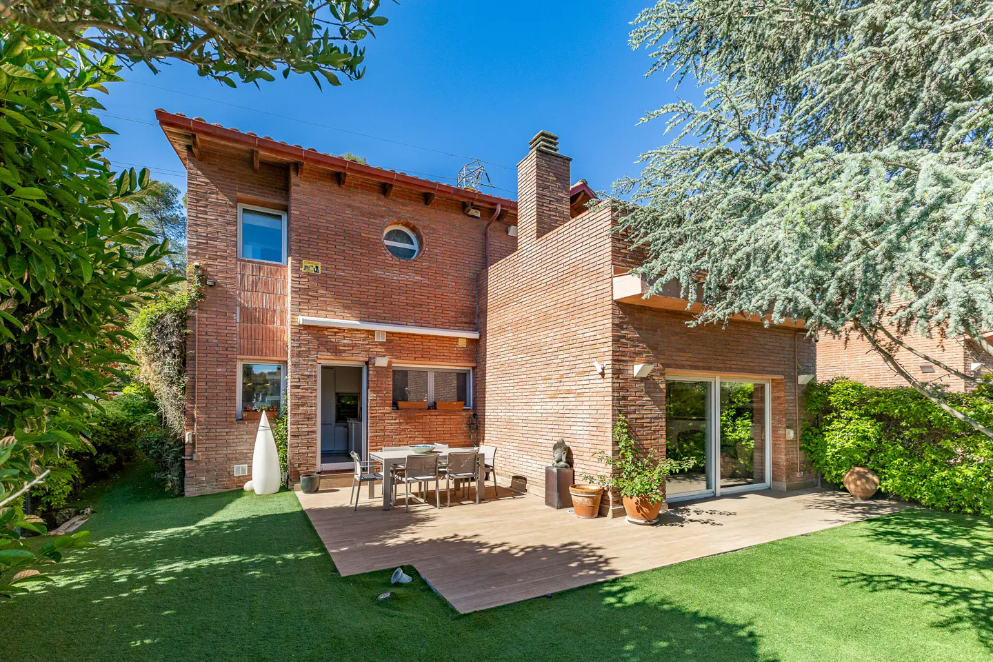 Backyard view of a two-story brick house with a chimney, patio, table, chairs, and green lawn under a clear blue sky.