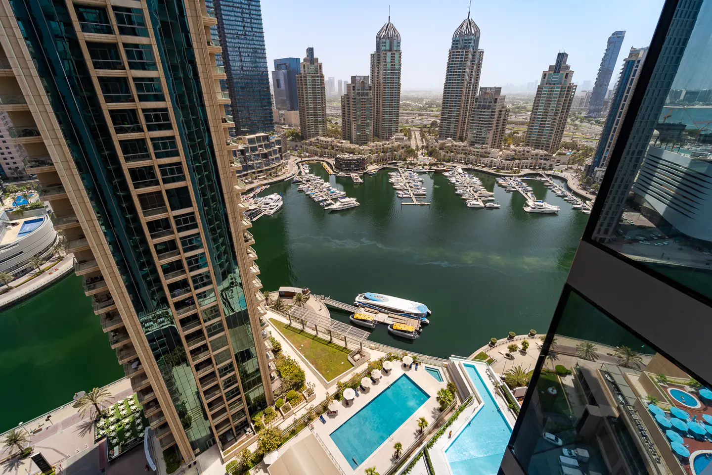 Aerial view of Dubai Marina with yachts, skyscrapers, and a blue swimming pool. Tall buildings frame the scene.