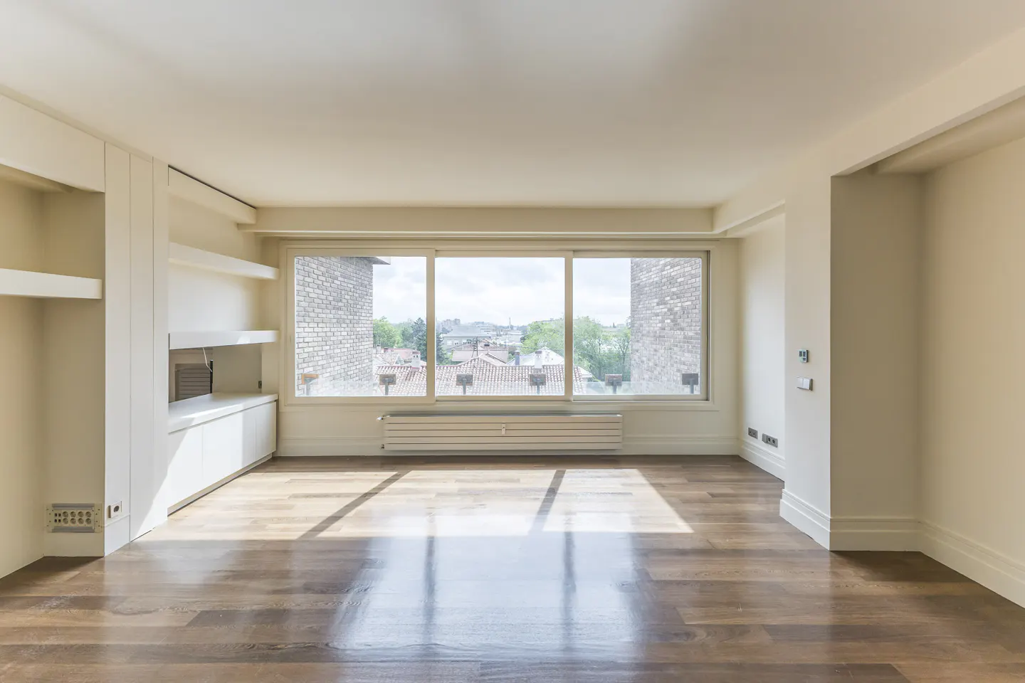 Bright, empty room with hardwood floors and a large window showing a cityscape. Built-in shelves and cabinets are on the left wall.