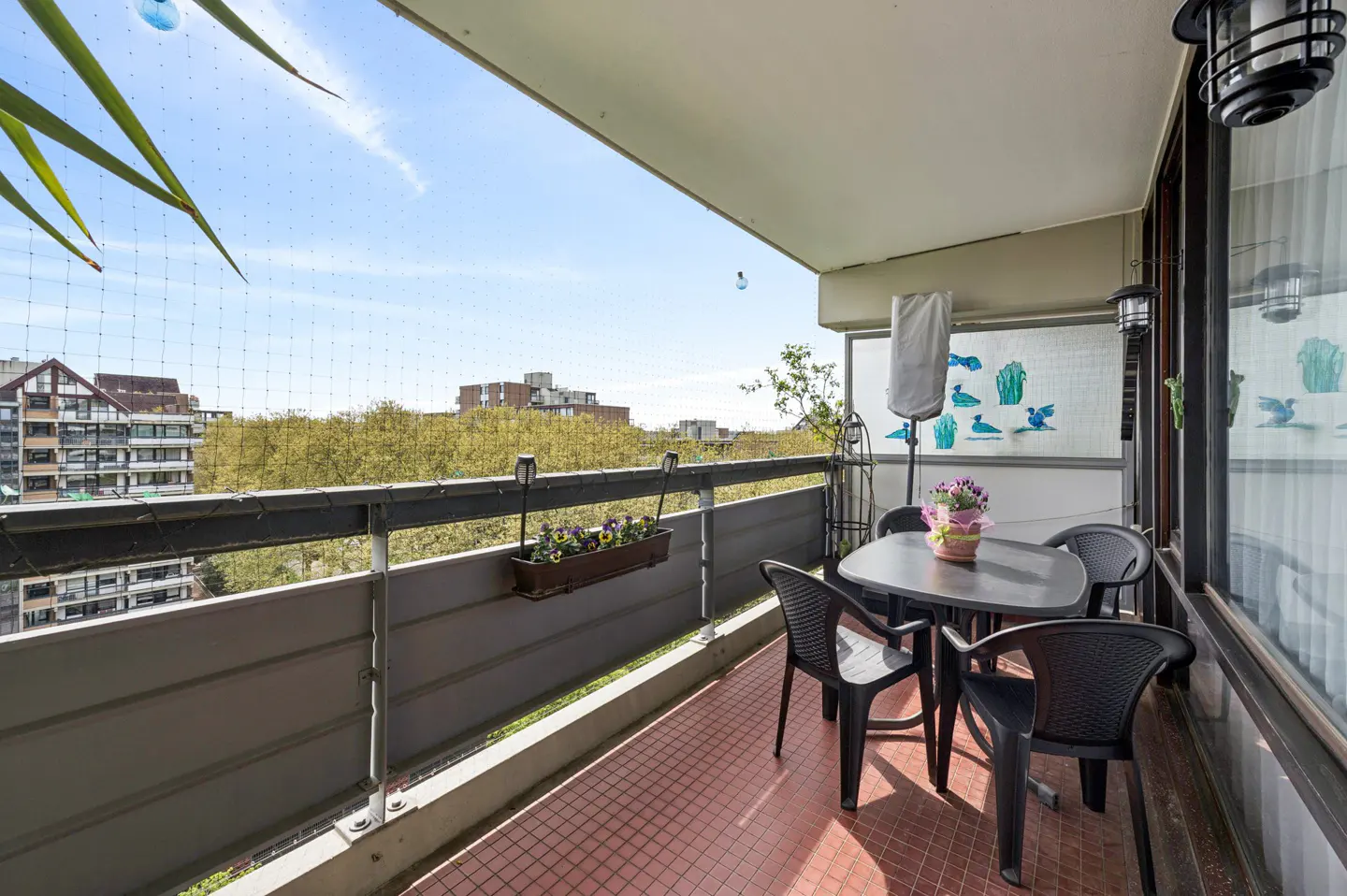 A balcony with a table and chairs overlooks a cityscape. A flower box hangs on the railing. The floor is tiled in red.