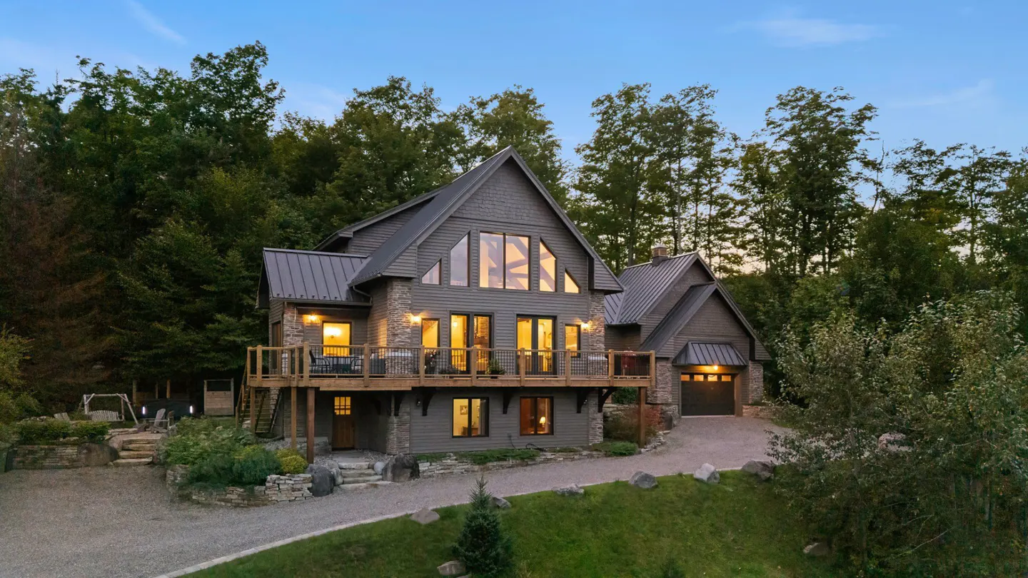 Two-story gray house with stone accents, a metal roof, and a wooden deck, surrounded by trees.