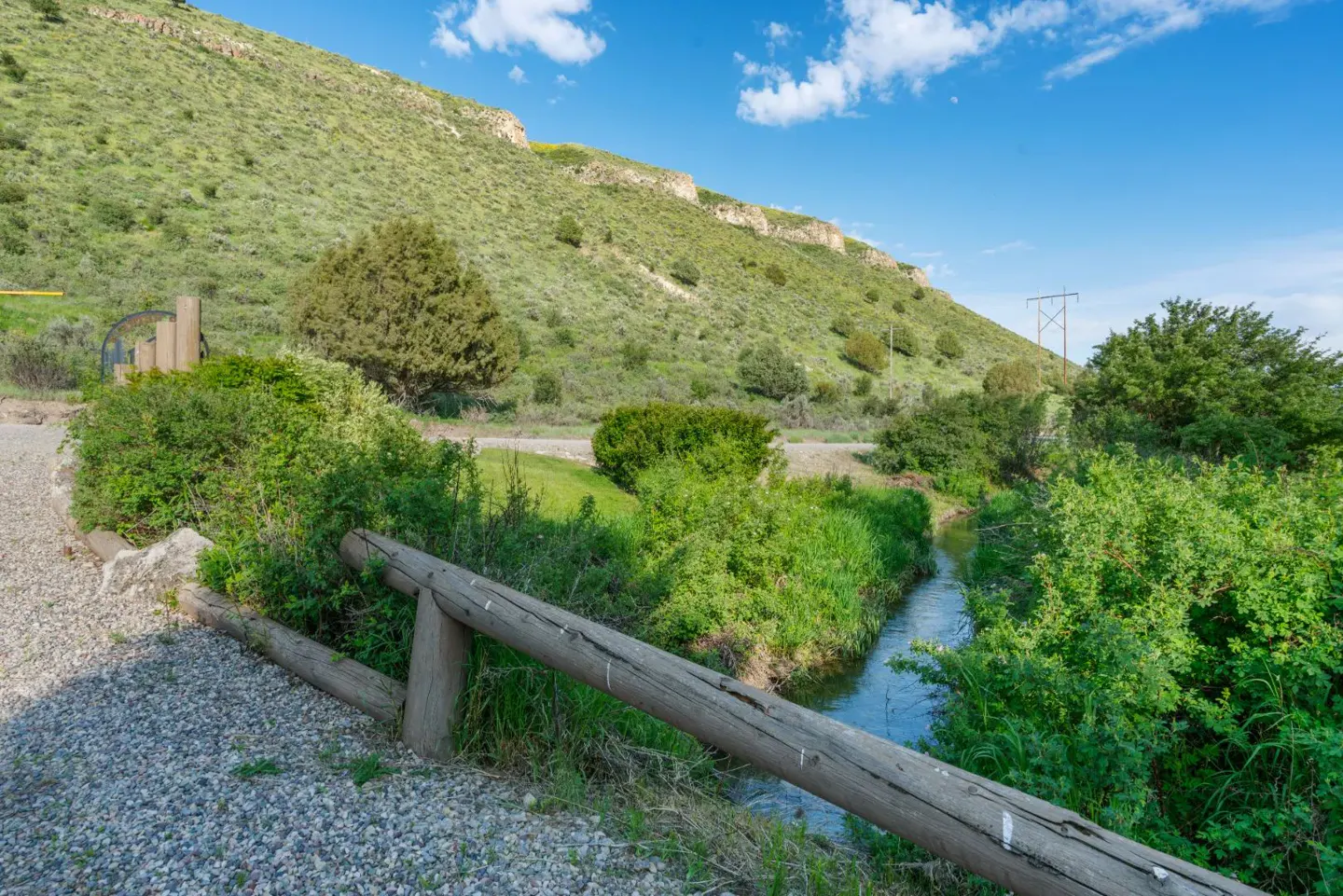 A small stream flows past a wooden fence next to a gravel path, with a large green hillside under a blue sky.