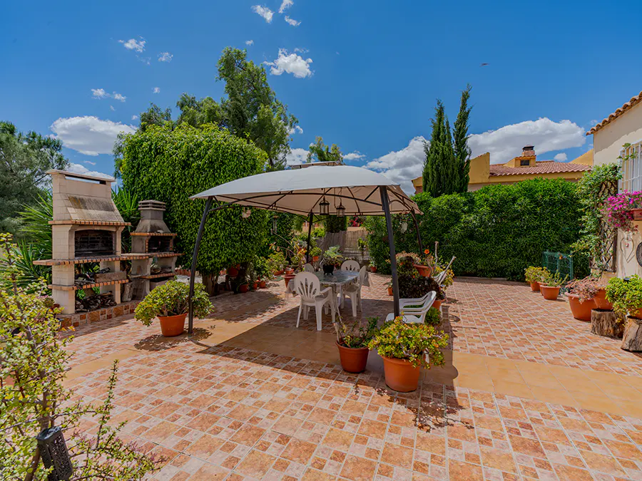 Outdoor patio with a white gazebo, table, and chairs on a tiled surface. Lush green plants and a brick barbecue are visible.