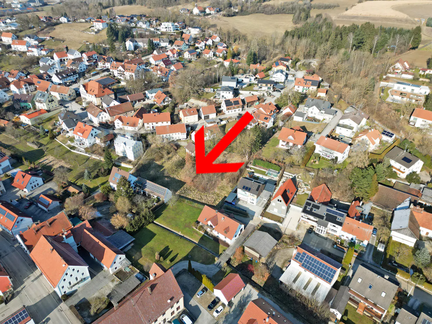 Aerial view of a residential area with an empty lot marked by a red arrow. Houses have red roofs and solar panels.