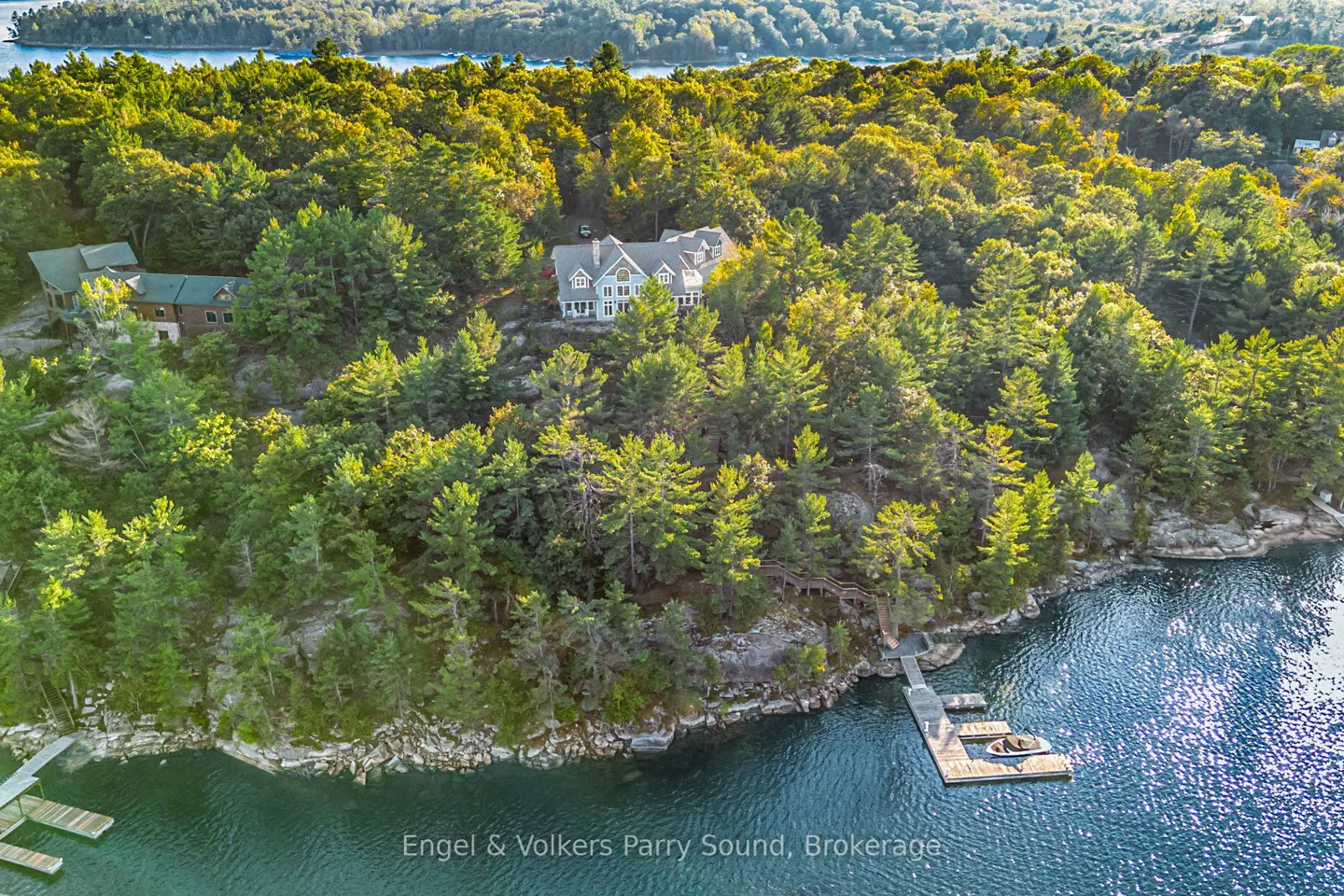 Aerial view of a grey house nestled in green trees on a rocky island with a dock on blue water.