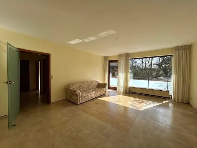 Bright, empty living room with beige walls, marble floor, floral sofa, and large window with white curtains.