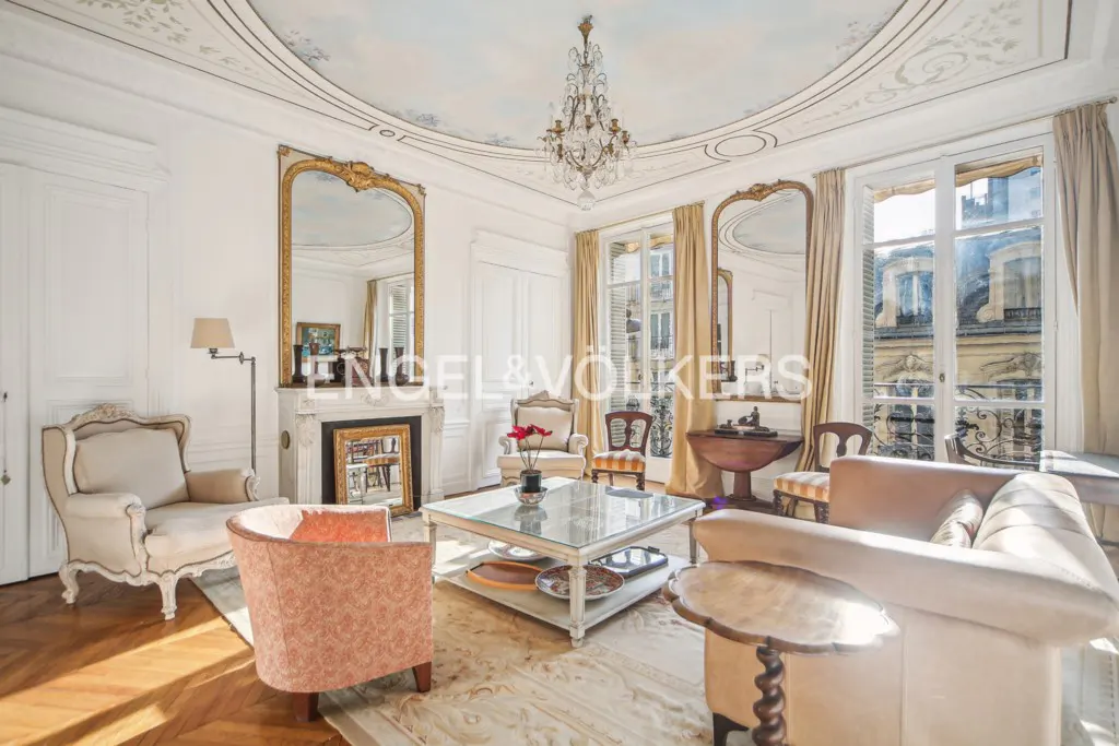 Elegant Parisian living room with a crystal chandelier, ornate fireplace, mirrors, and classic furniture on wood floors.