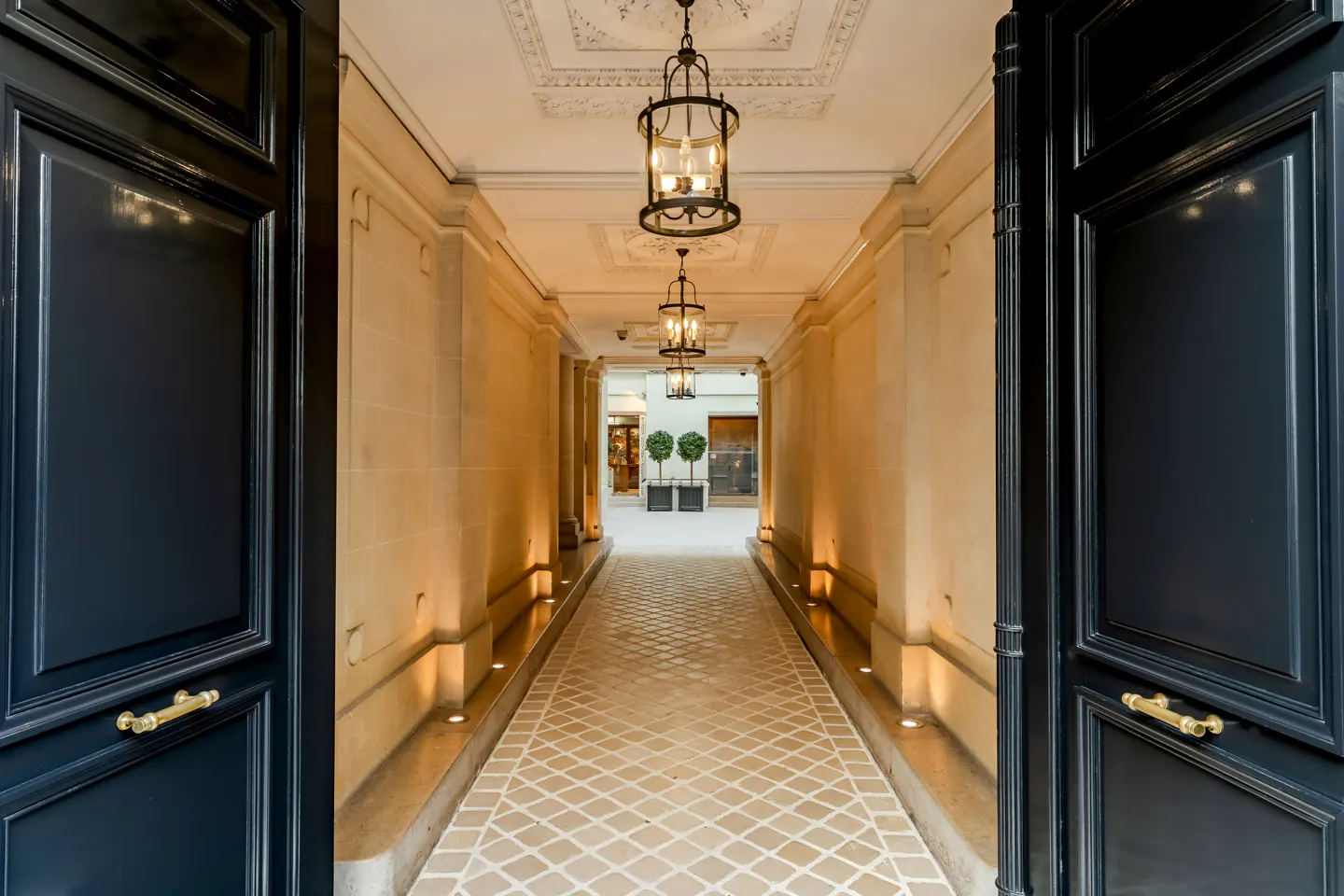 View through open dark blue doors into a lit stone hallway with hanging lanterns and a patterned floor leading to a courtyard.