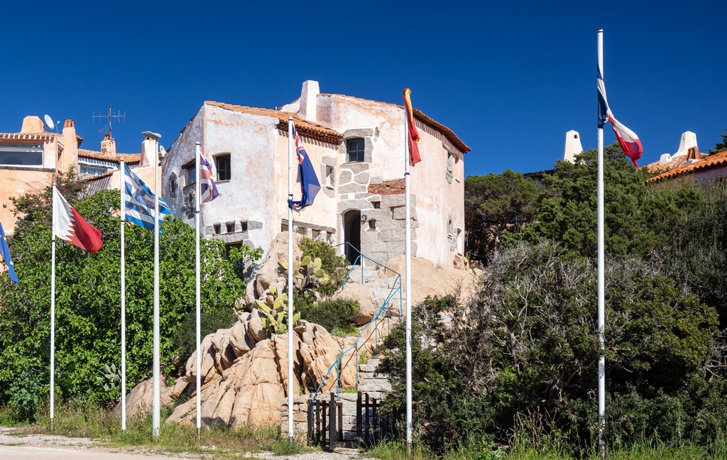 Exterior view of a stucco house on a rocky hill with flags of different countries in front.