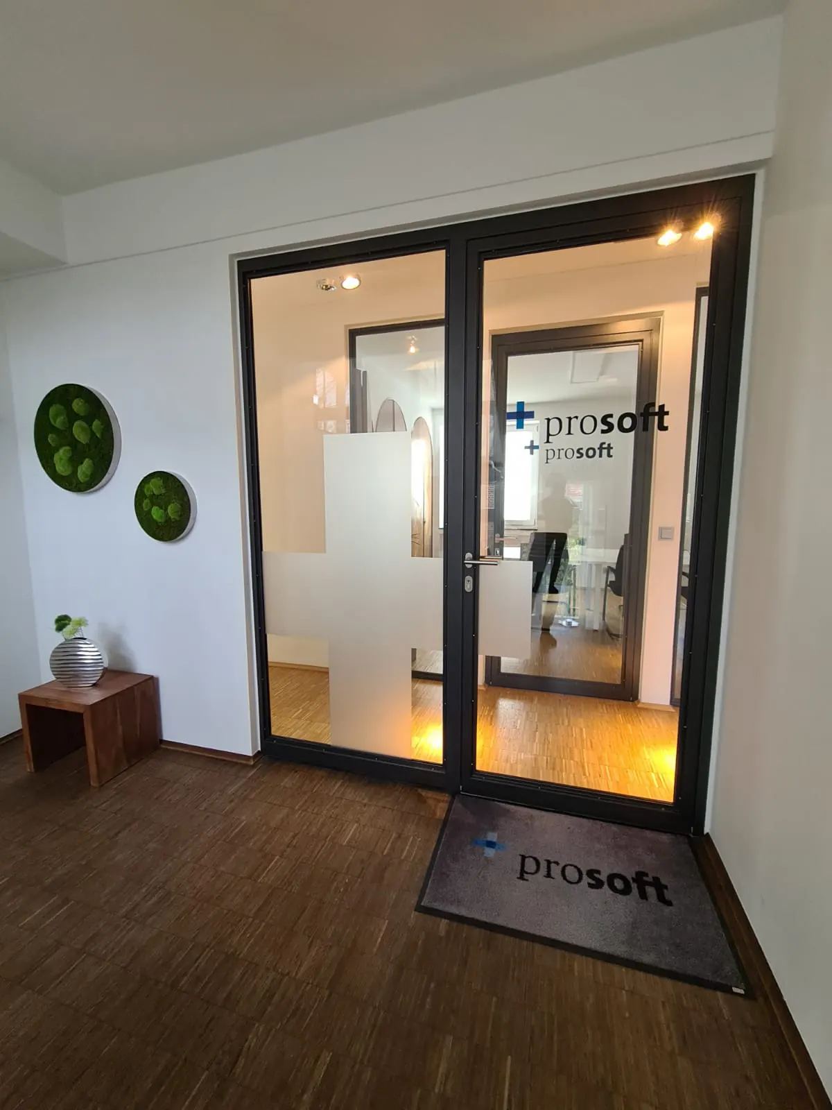 Office entrance with glass doors, "prosoft" logo, and a cross design. Wooden floor, moss wall art, and a small table with a plant.