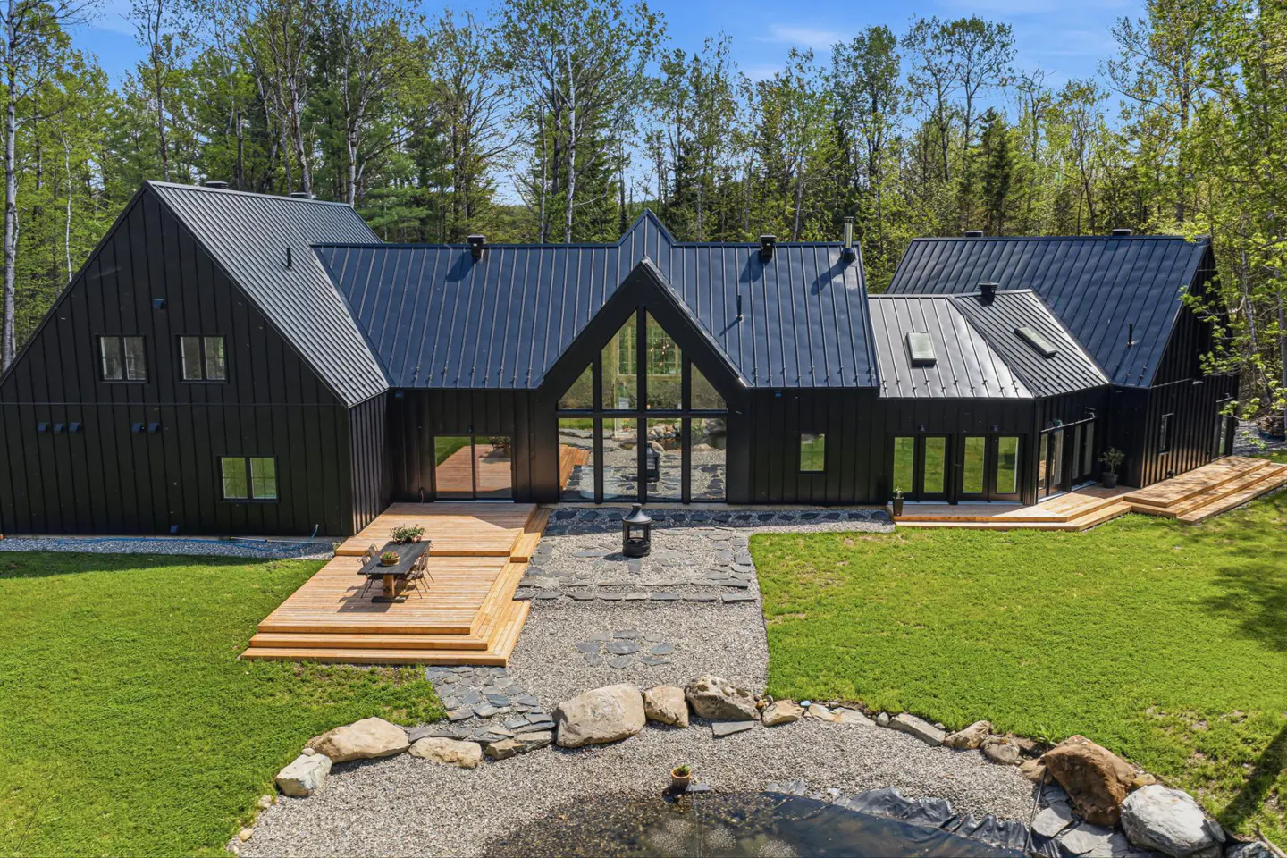 Aerial view of a modern black house with a metal roof, wooden deck, and a pond in the front yard.
