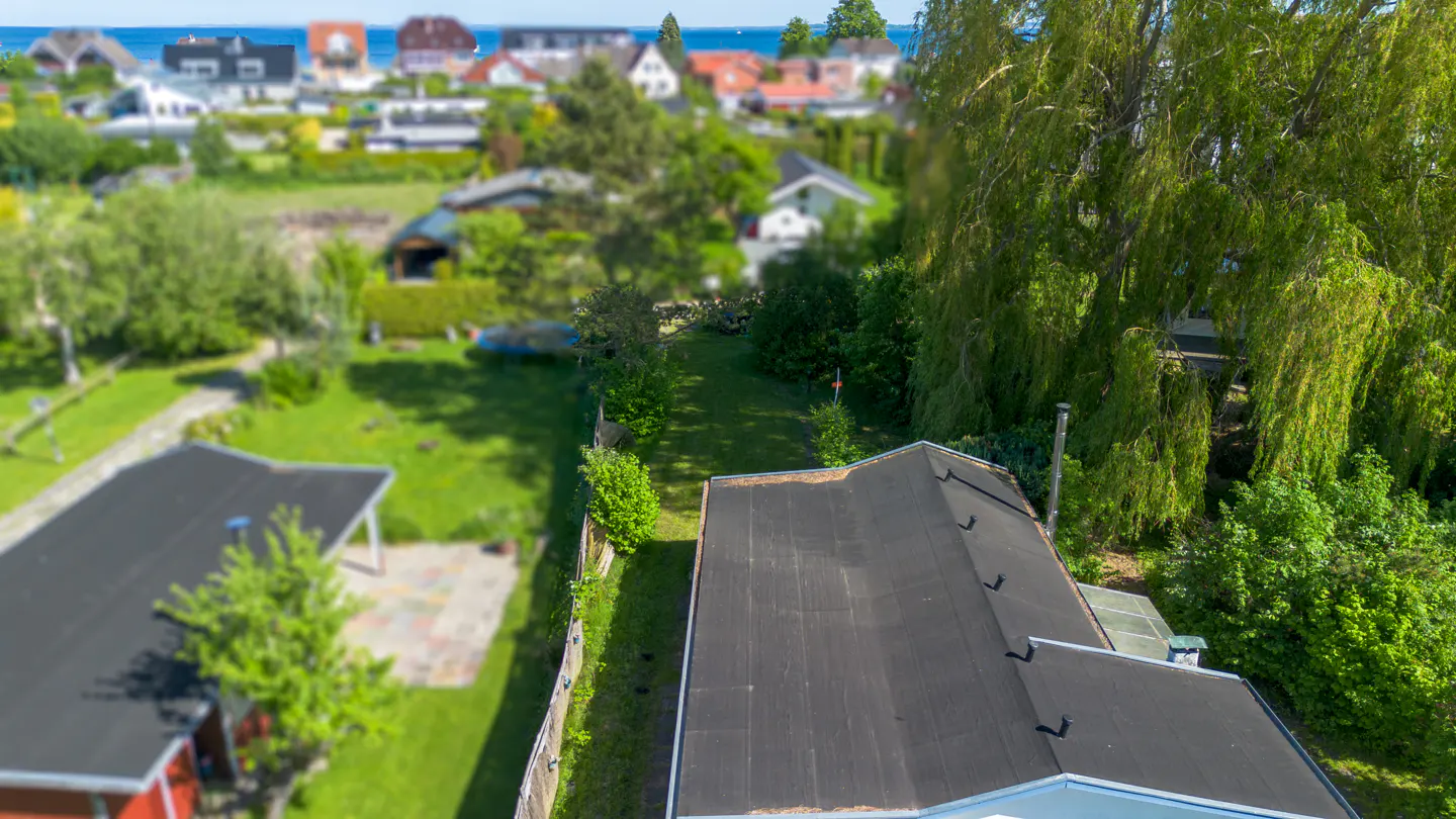 Aerial view of a house with a black roof, surrounded by lush green trees and a lawn, with a coastal town in the distance.