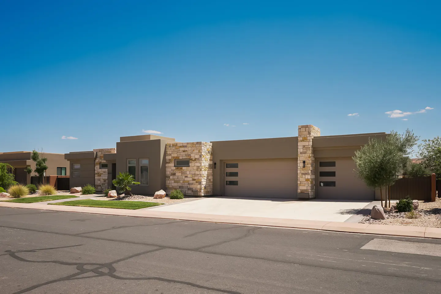 Modern, single-story homes with tan stucco and stone accents under a clear blue sky. Driveways lead to attached garages.