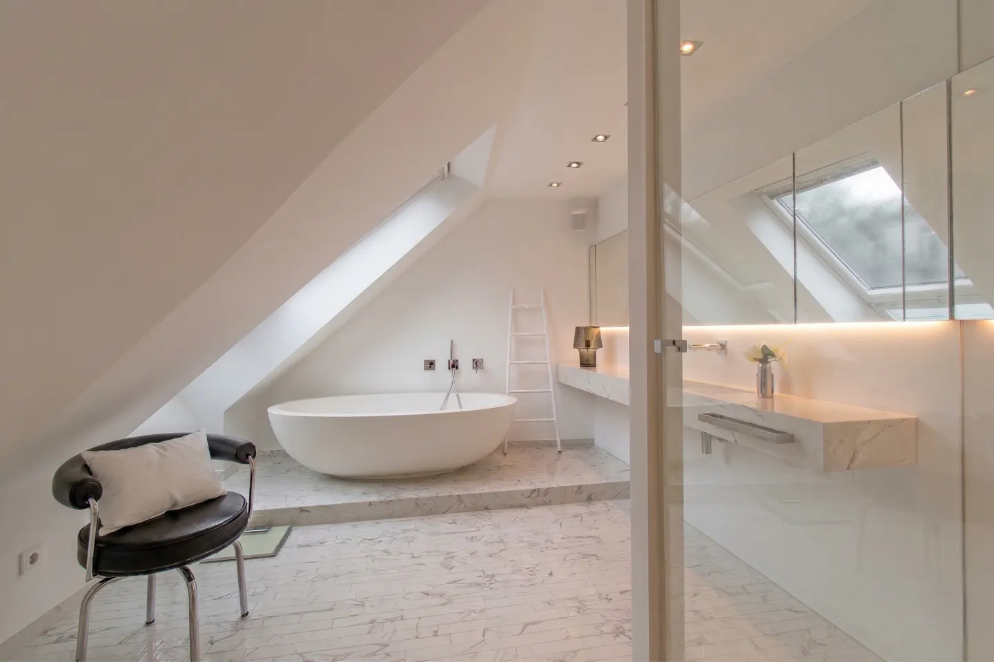 Bright, modern bathroom with white walls, marble floor, and a freestanding tub under a skylight. A black chair sits in the foreground.