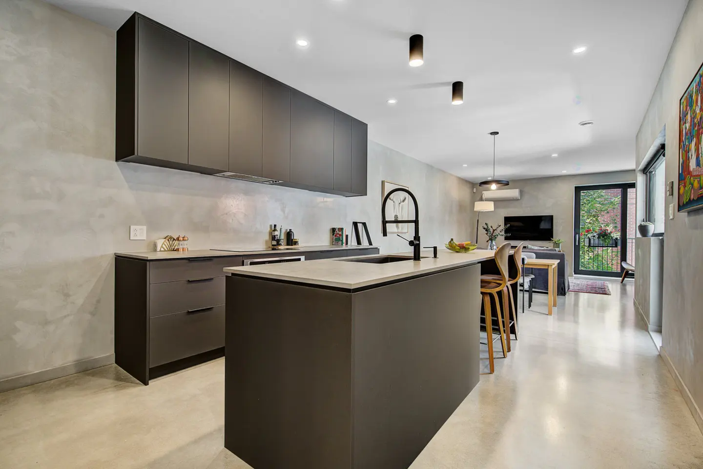 Modern kitchen with gray cabinets, island, and concrete floors. Living area visible in the background.
