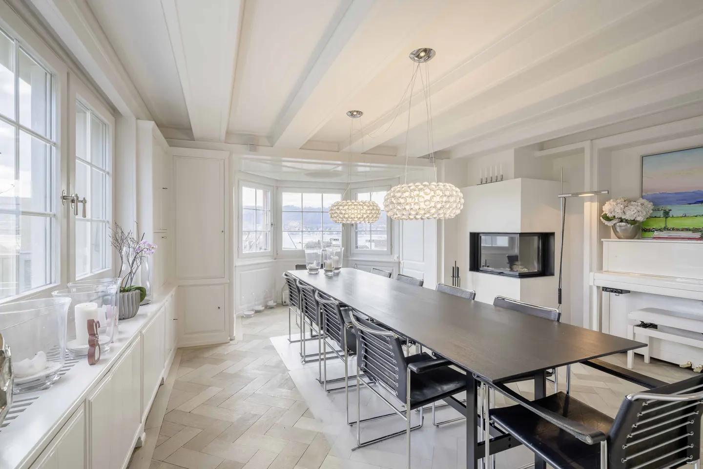 Bright dining room with a long black table, black chairs, and three crystal chandeliers. A white piano and fireplace are in the background.
