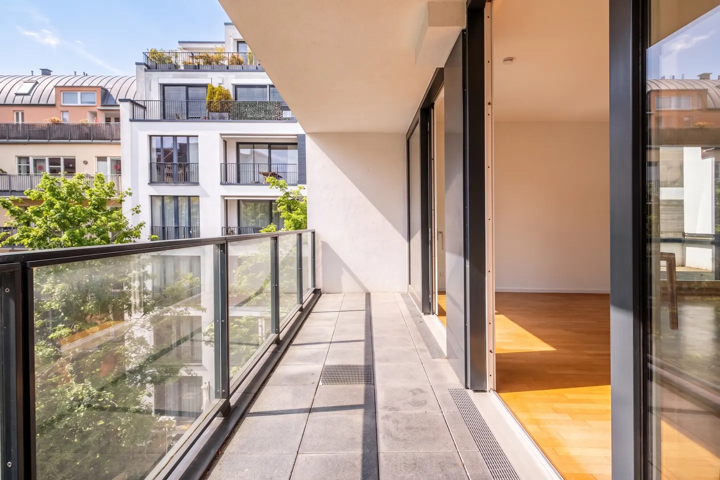 A modern apartment balcony with glass railings and gray tiles, leading into a bright room with hardwood floors.