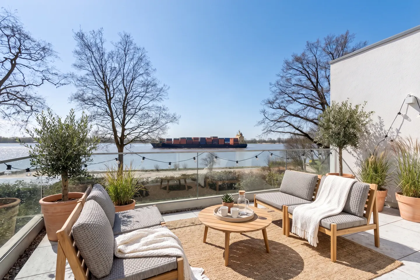 Outdoor patio with wood furniture, a round table, and a view of a river with a cargo ship on a sunny day.