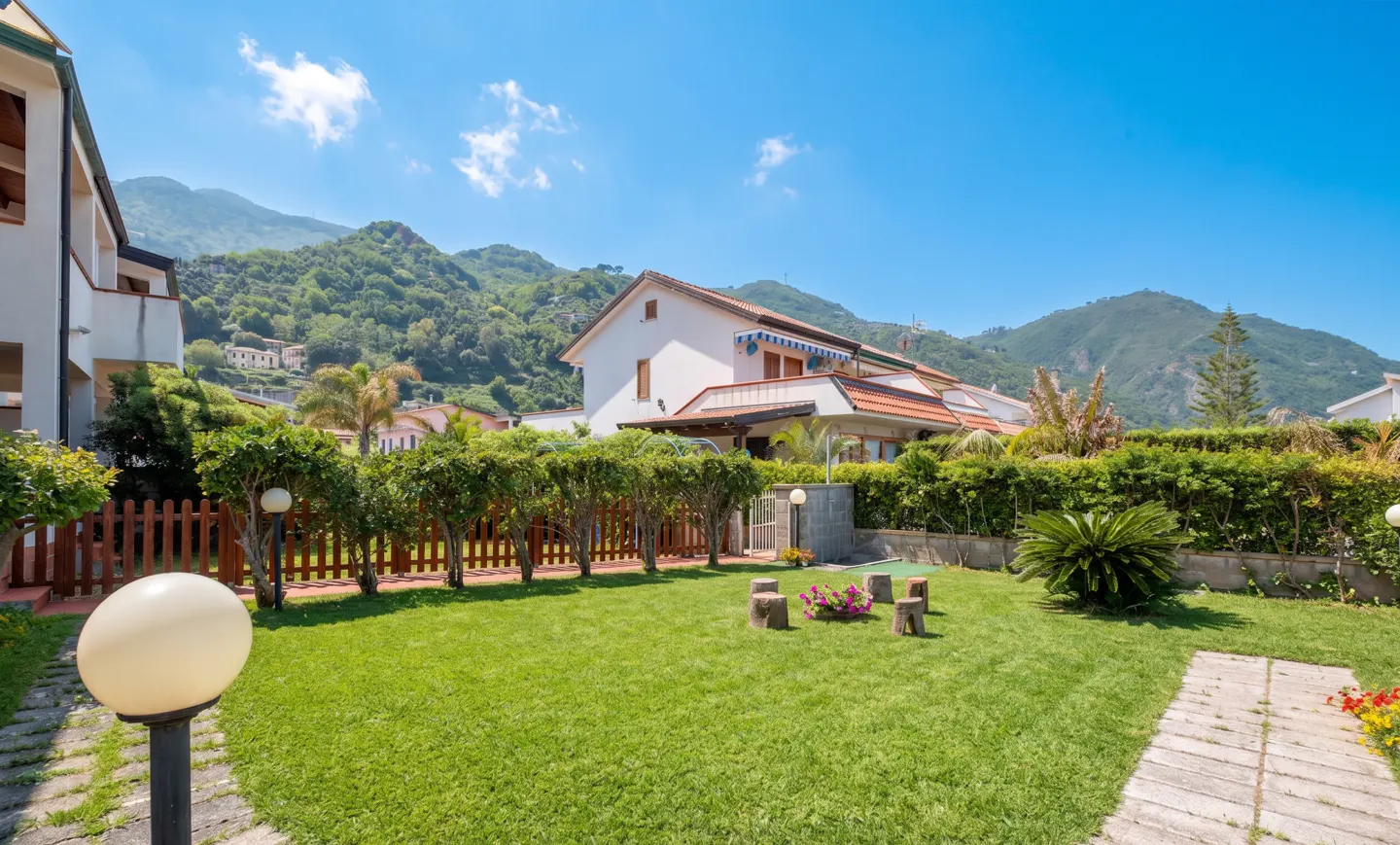 Lush green lawn with a wooden fence, hedges, and a white house with a red tile roof under a blue sky with mountains in the background.