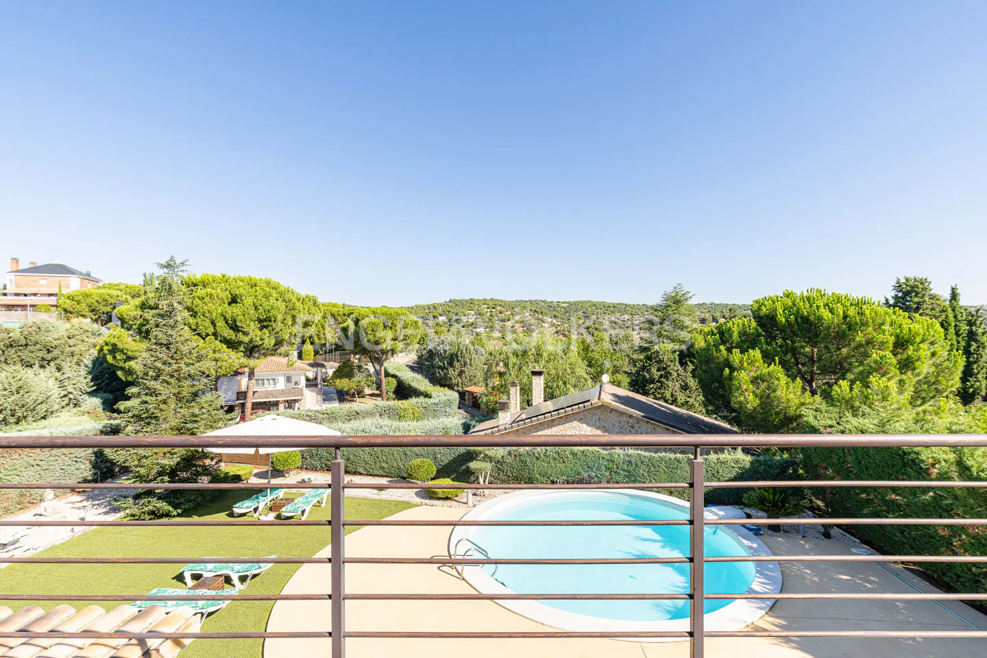 View from balcony of a blue pool, green lawn, and lounge chairs. Houses and trees are in the background under a clear blue sky.