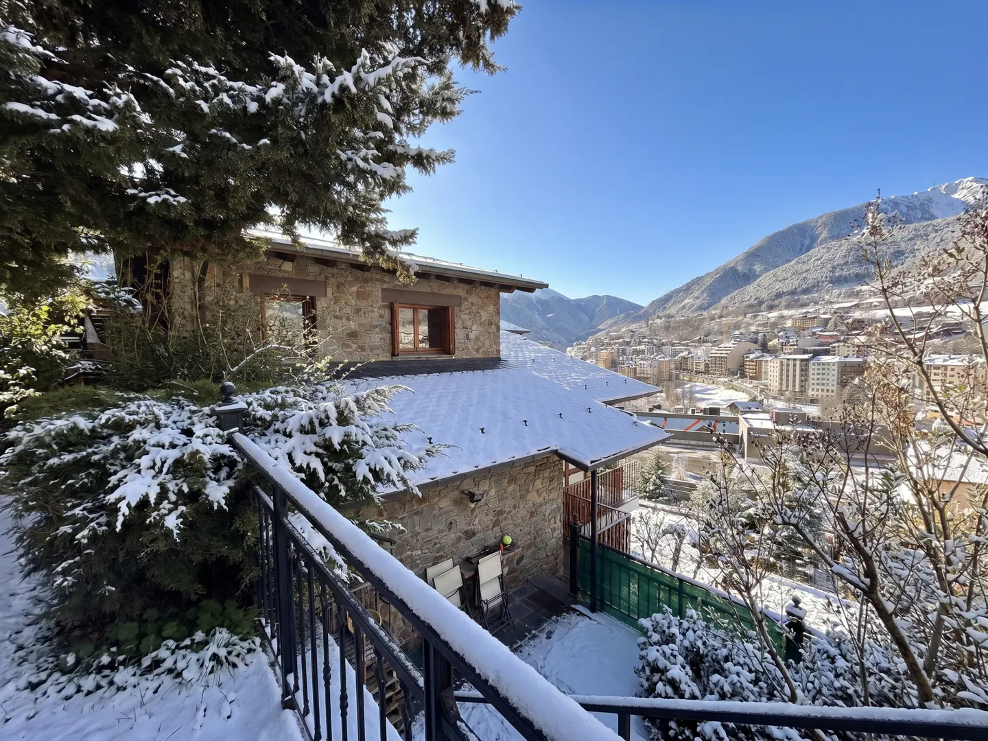 A stone house with a snow-covered roof overlooks a valley town on a sunny day.