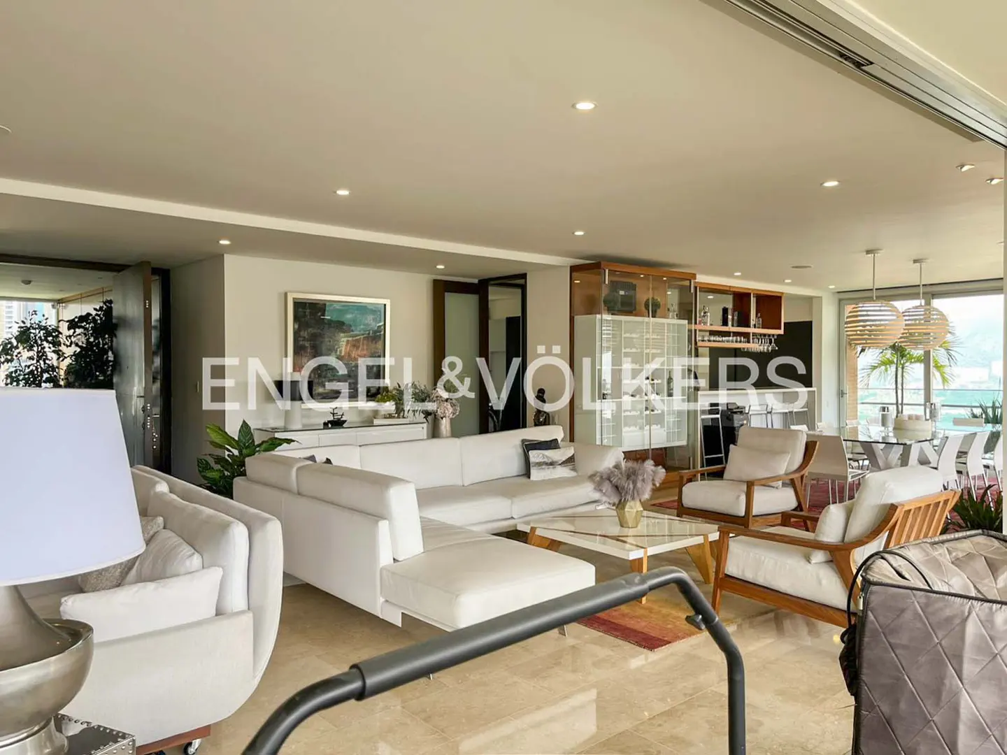 Bright living room with white sofas, armchairs, and a glass coffee table. A bar area and dining table are visible in the background.