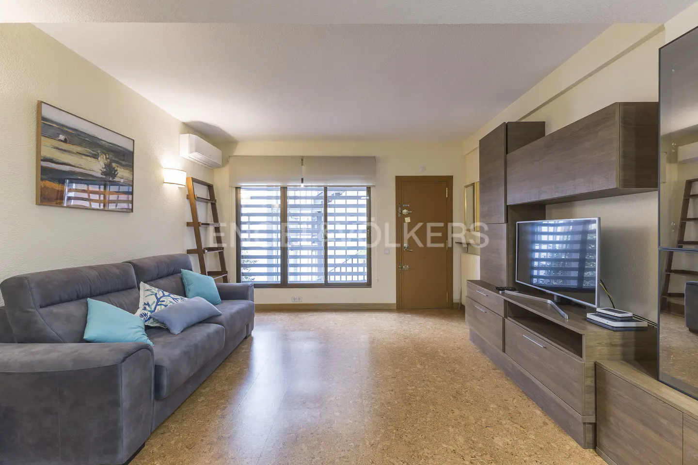 Living room with gray sofa, blue pillows, wood floors, and a large window. A TV and shelving unit are on the right wall.