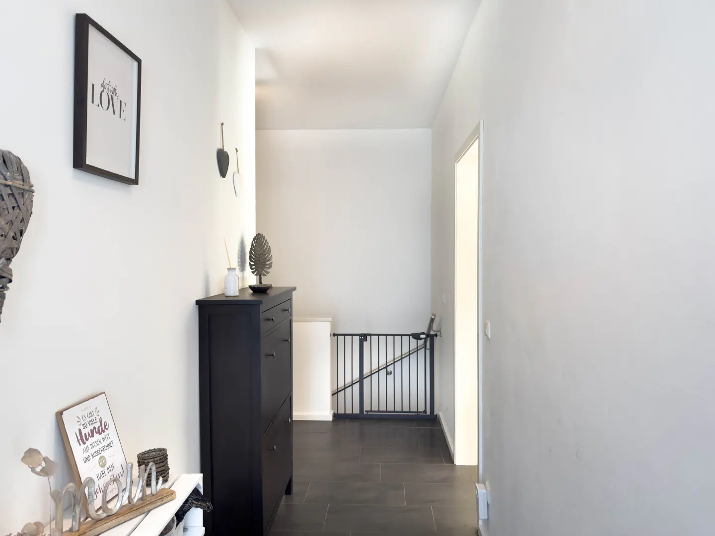 Hallway with white walls, dark tile floor, and black furniture. A baby gate blocks the stairs. A framed "LOVE" print hangs on the wall.