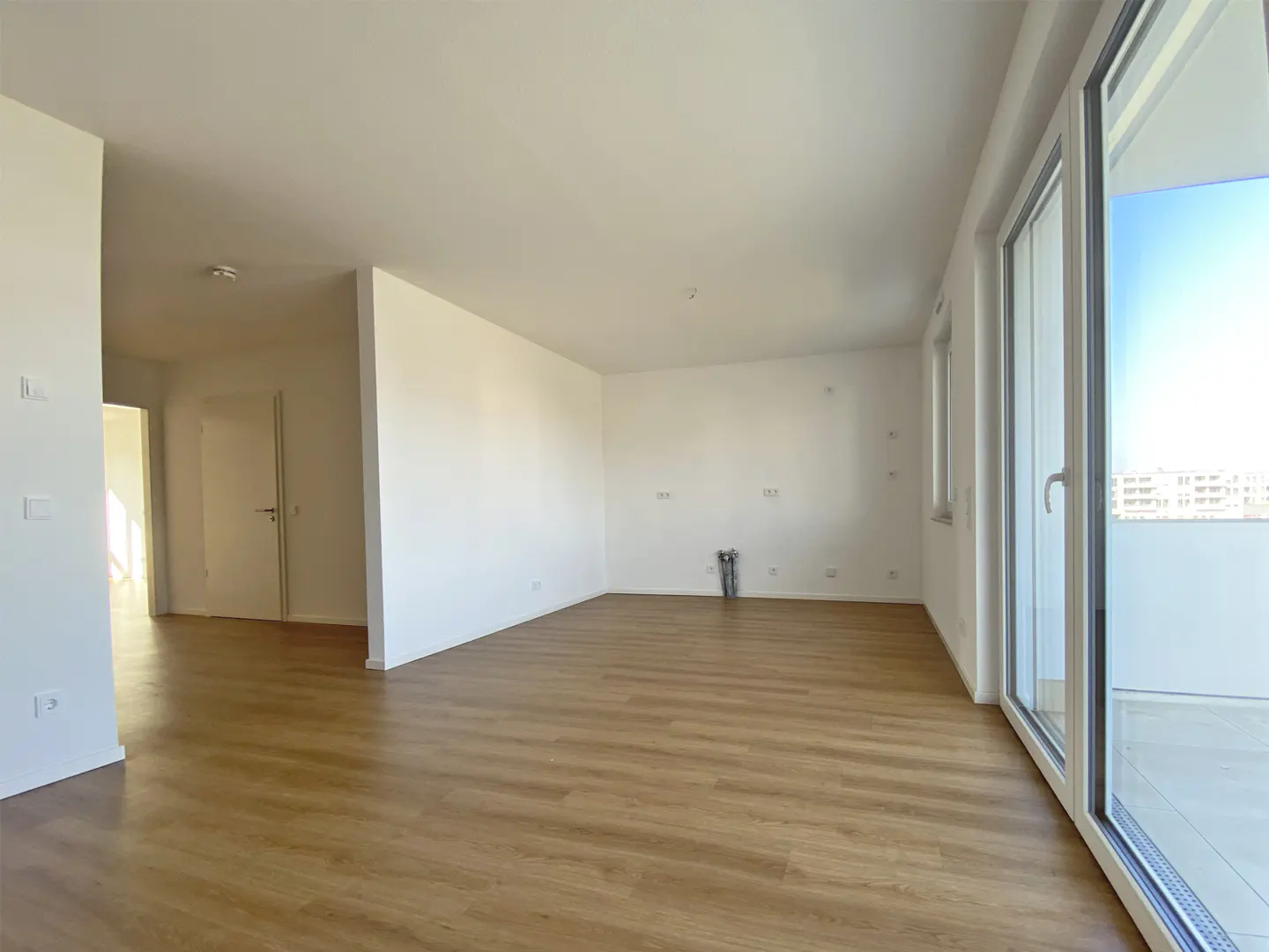 Bright, empty room with light wood floors and white walls. Large sliding glass doors lead to a balcony with a city view.