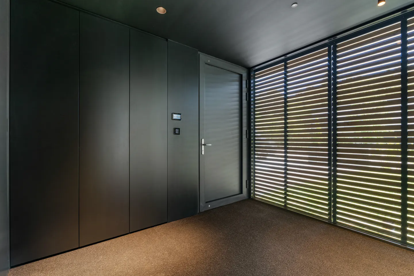 Modern entryway with matte black wall panels, a matching door, and large windows covered by horizontal wooden blinds.