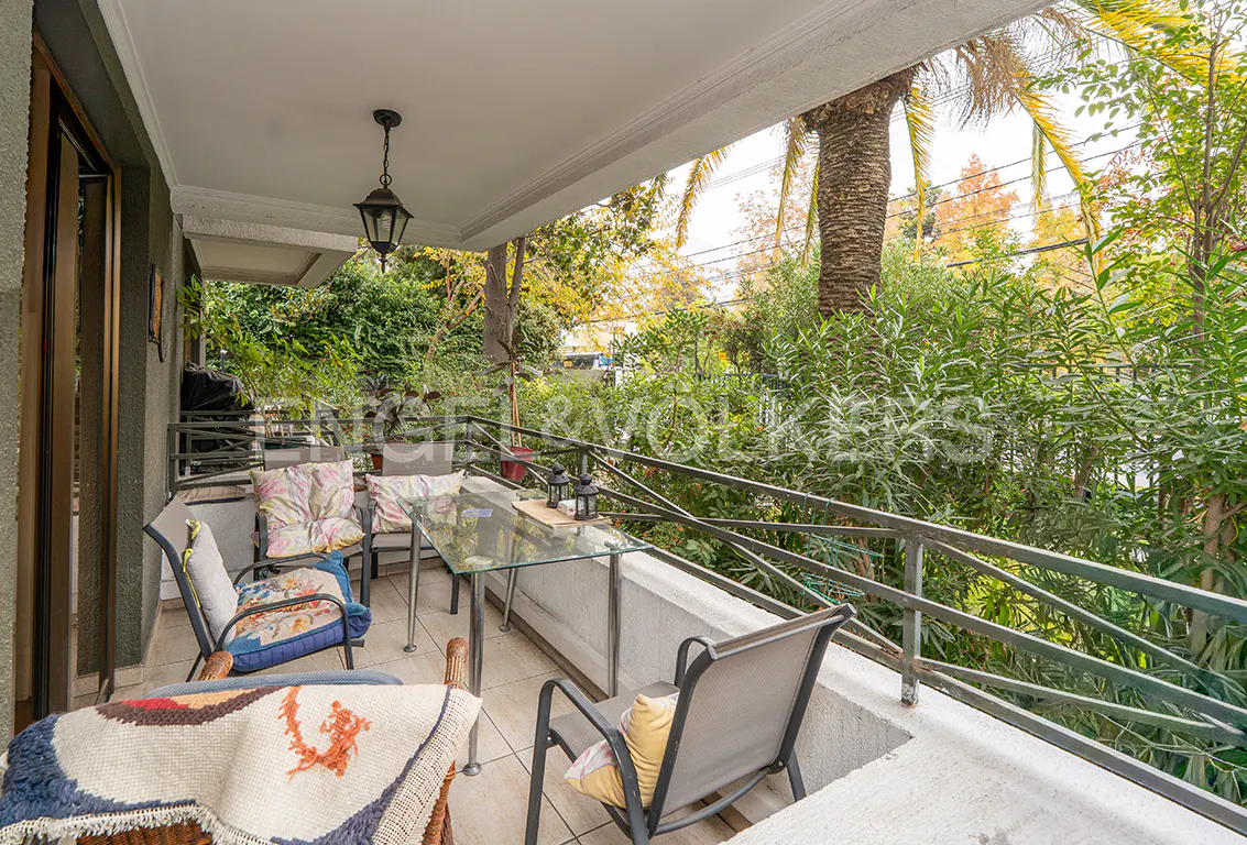 A covered patio with a glass table, chairs, and a view of lush greenery. A hanging lantern adds a cozy touch.