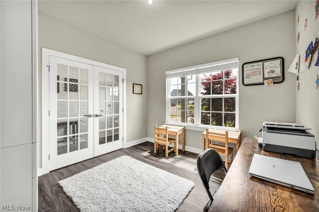 Bright home office with wood floors, white walls, and a large window. A desk with a laptop and printer sits near two small wooden desks.