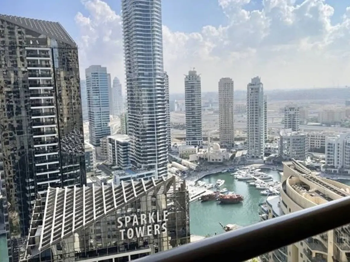 View from a balcony overlooking Dubai Marina, featuring the Sparkle Towers and numerous skyscrapers. Boats are docked in the turquoise water.