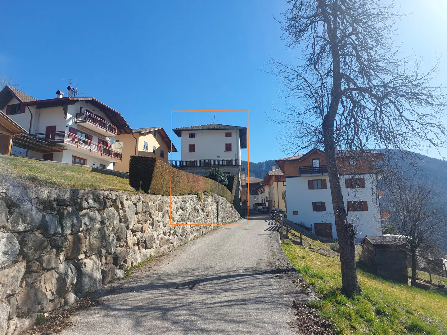 A white, two-story house with red shutters sits above a stone wall on a sunny day. A narrow road leads up to the house.