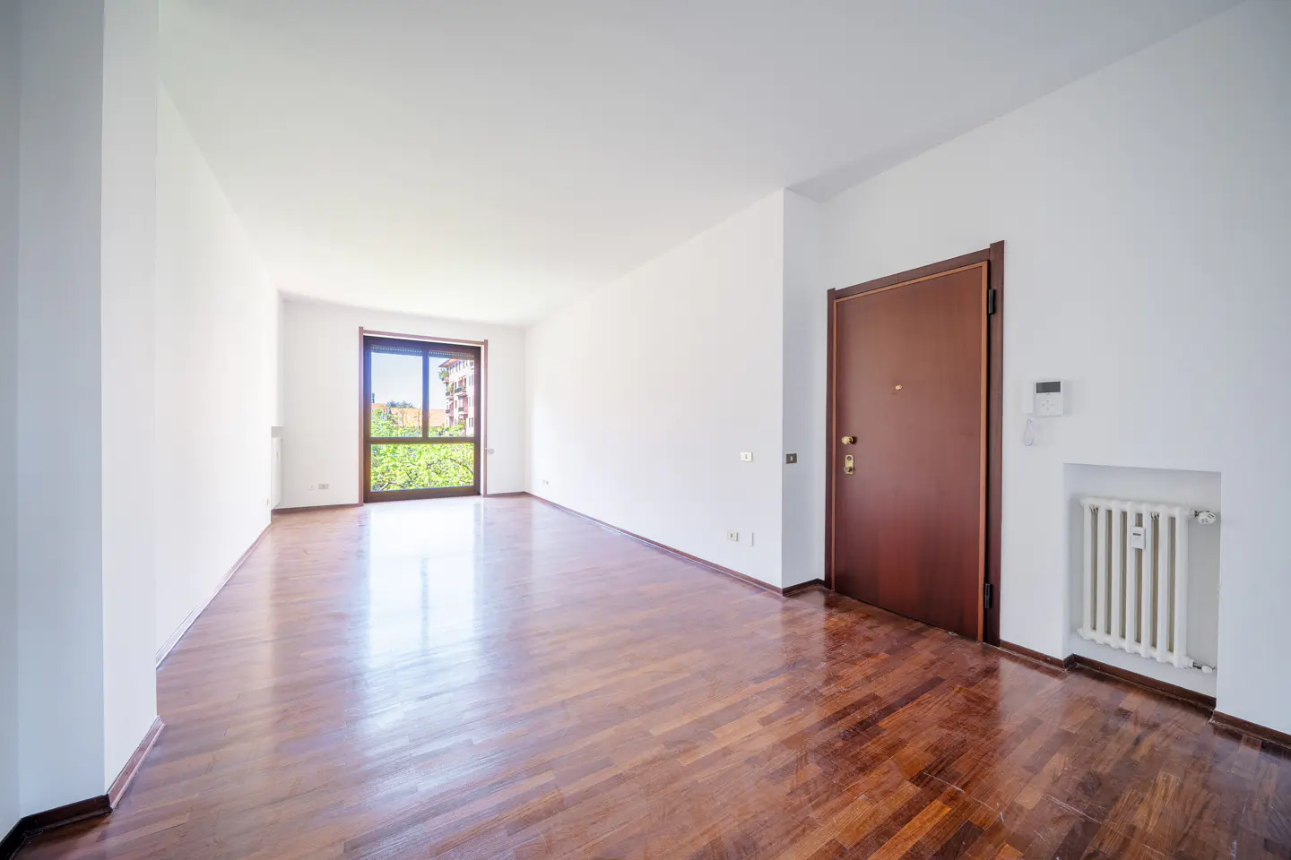 Bright, empty room with white walls, dark wood floors, and a brown door. A window shows green trees outside. A white radiator is on the wall.