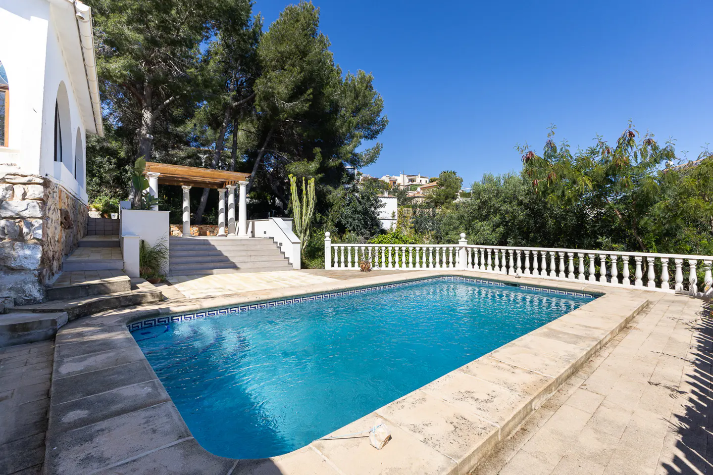 A rectangular swimming pool with turquoise water, surrounded by stone tiles, sits in a backyard with trees and a pergola.