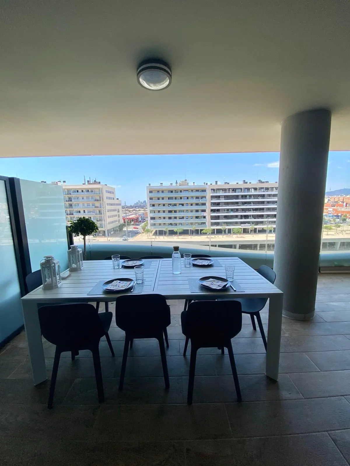 Covered balcony with a white dining table, black chairs, and a scenic view of modern apartment buildings under a blue sky.