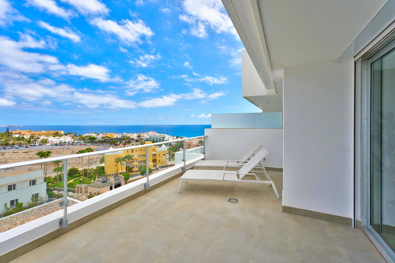 Modern balcony with two white lounge chairs overlooking a coastal town and the blue ocean under a sunny, cloudy sky.