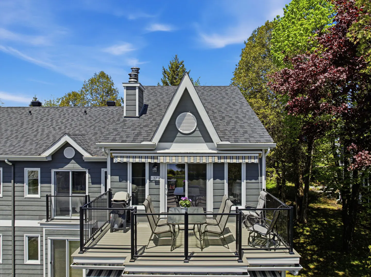 Exterior view of a gray house with a deck, table, chairs, grill, and trees against a blue sky.