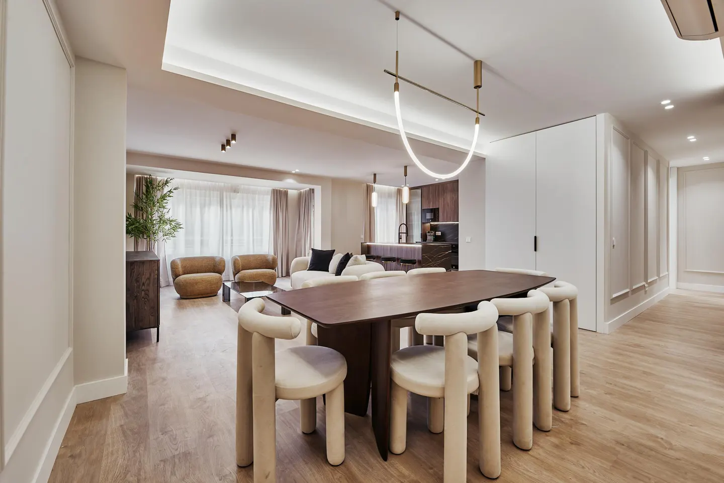 A modern, open-concept living and dining area with a dark wood table, cream chairs, and a sleek, curved light fixture.