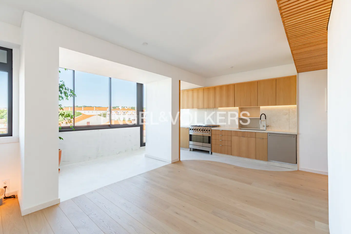 Bright, modern apartment with light wood floors, white walls, and a kitchen with wood cabinets. Large windows offer a city view.