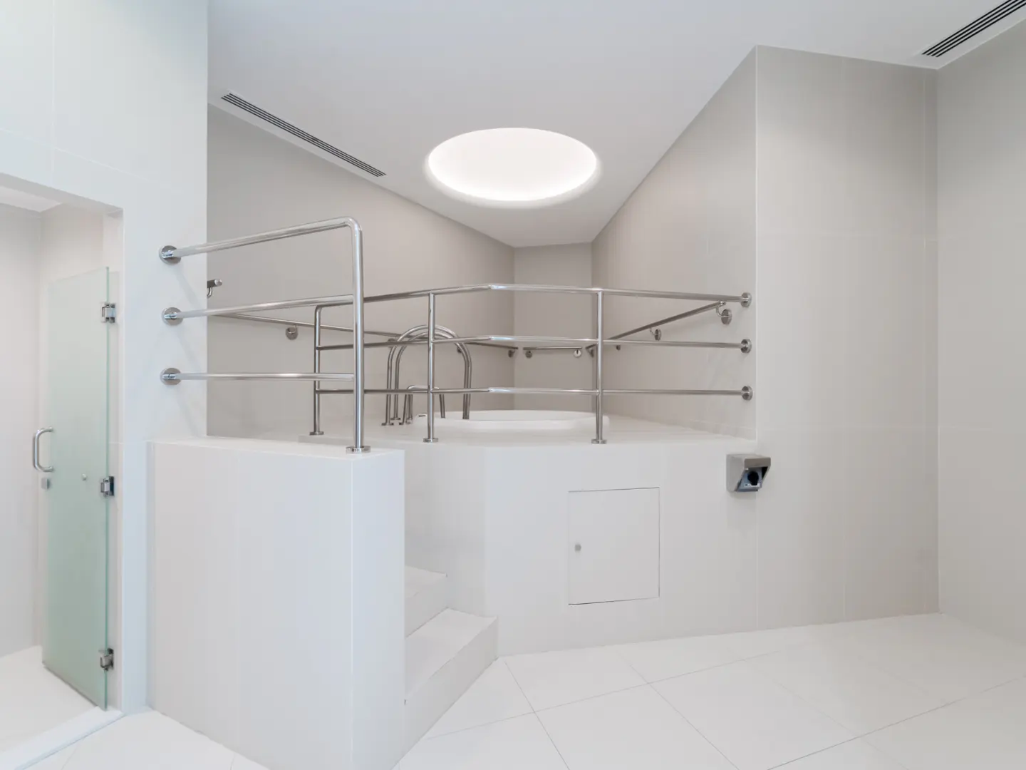 Minimalist white spa room with a raised circular tub, chrome safety railings, and a circular skylight above.