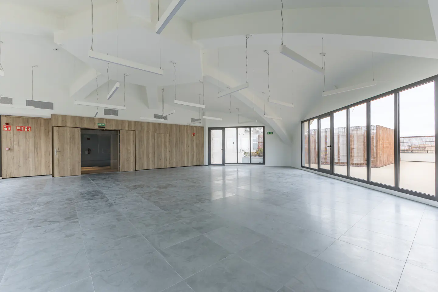 Bright, empty room with gray tile floor, white walls, and a high, peaked ceiling. Large windows line one wall, and wood-paneled doors are on another.