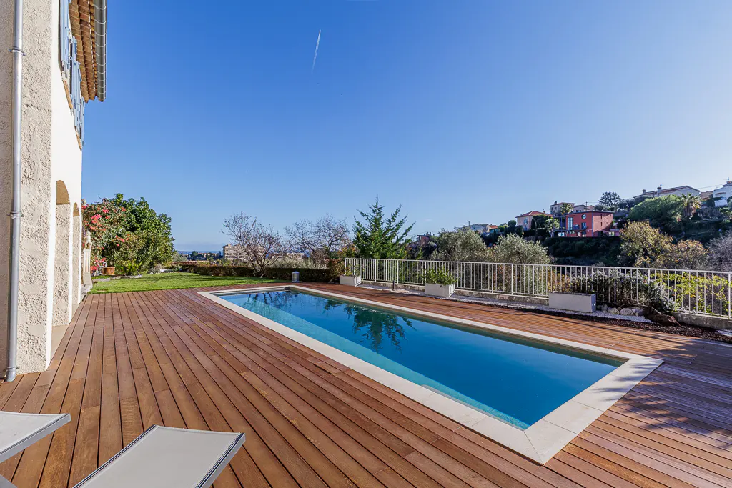 A rectangular pool with blue water is surrounded by a wooden deck and a white fence, with a view of houses on a hill.